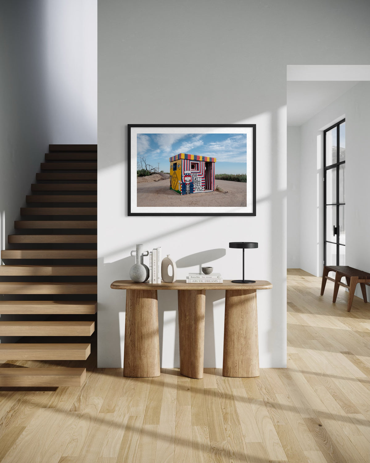 Wooden staircase in a sunlit hallway, “The Last Free Place” photo displayed above a console table.