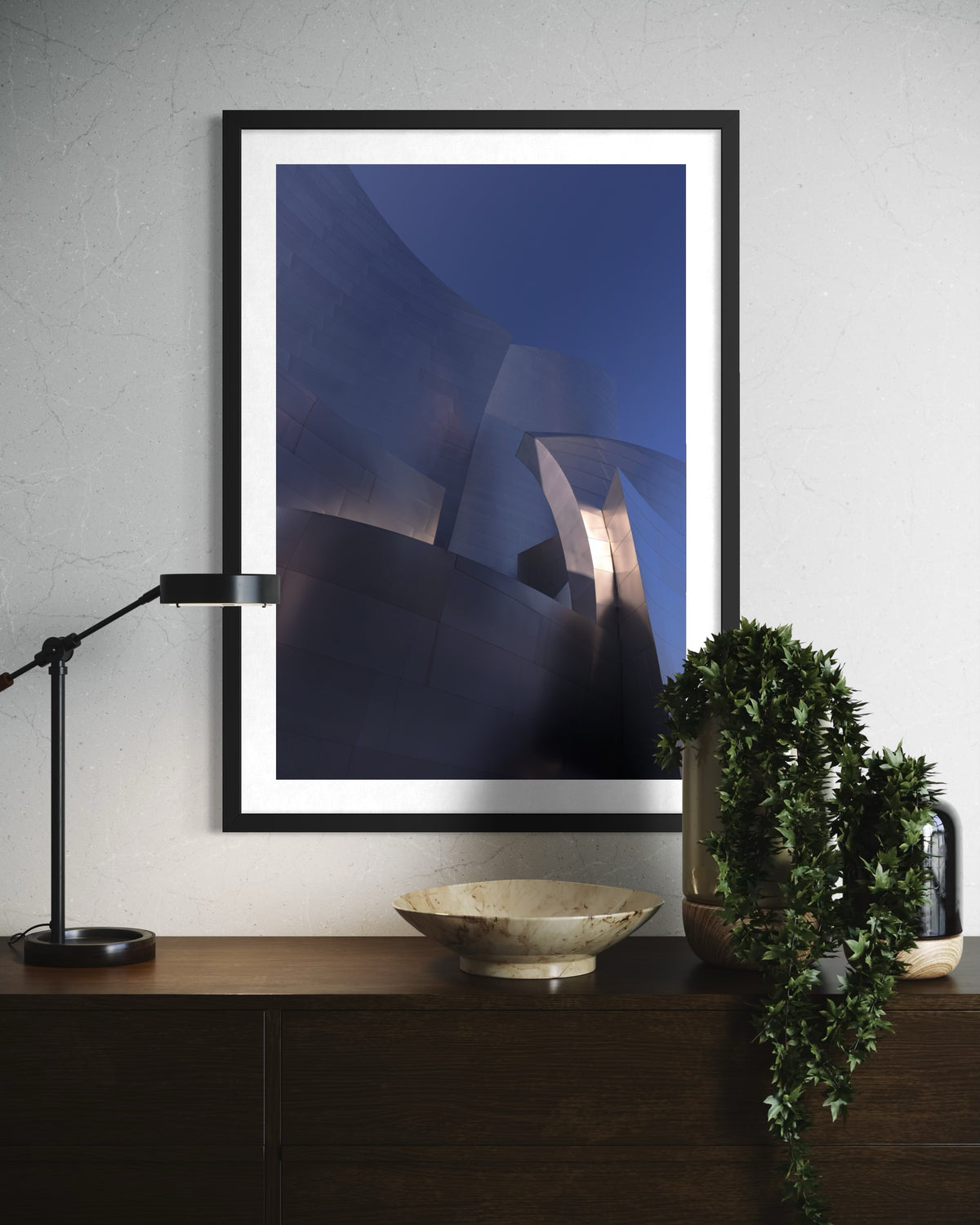 Abstract print of blue-tinted Frank Gehry architecture displayed above a sideboard with lamp, bowl, and plant.