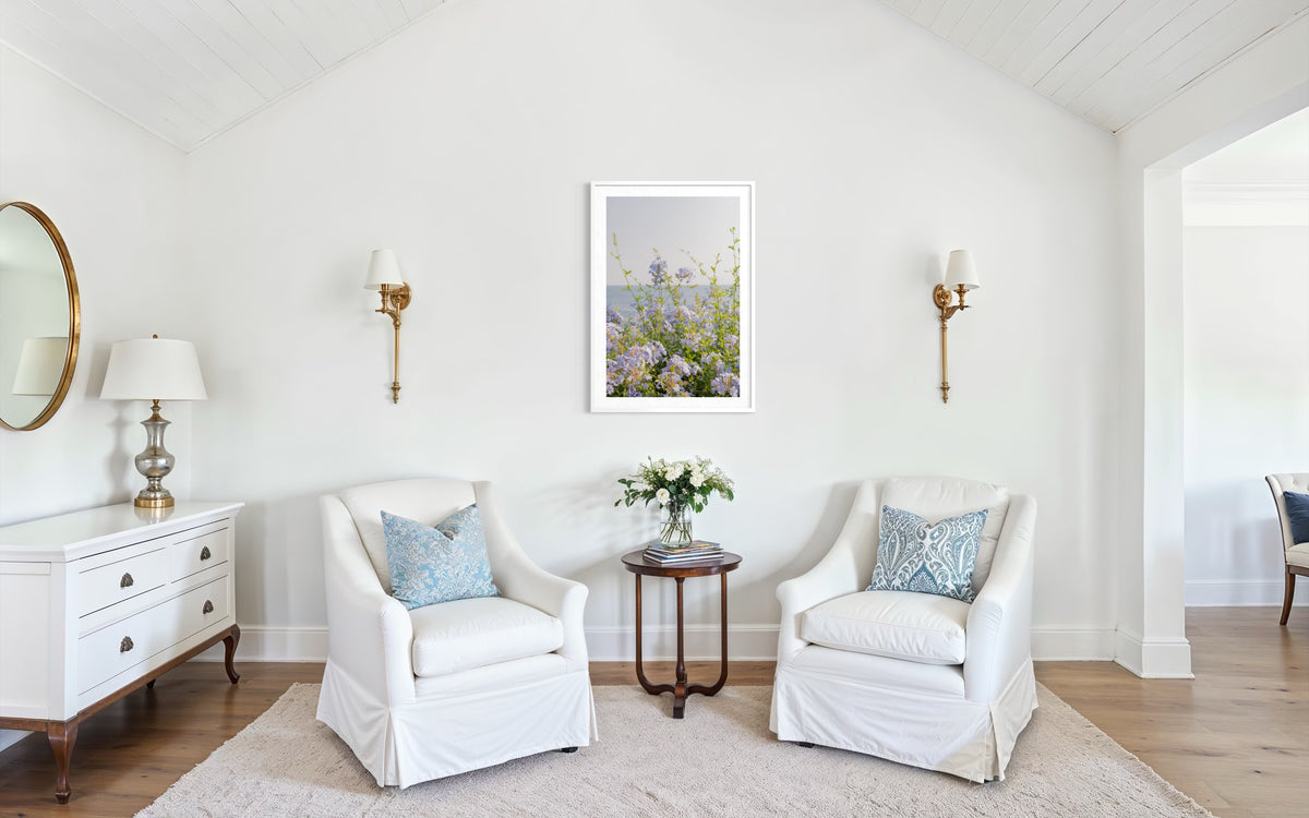 Two white armchairs with blue pillows face a round table and vase in a sunlit, elegant living room.