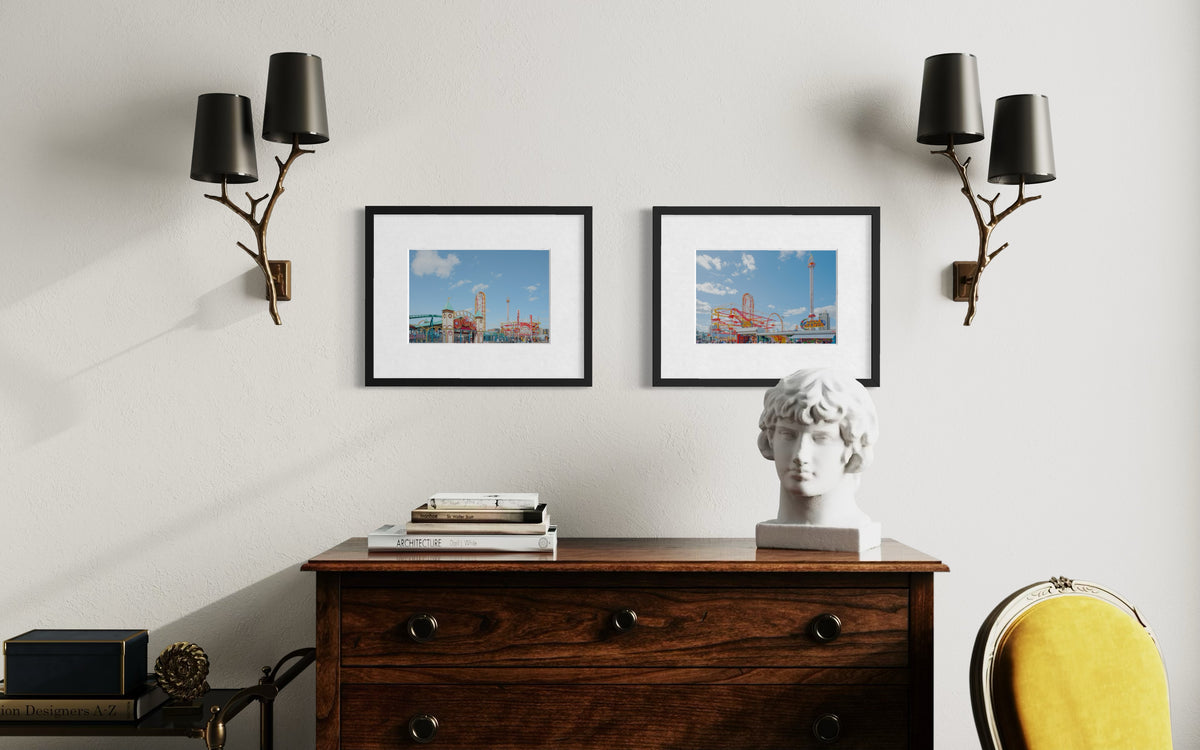 A dresser with books, a white bust, and framed Coney Island artwork beneath wall sconces in a home setting.