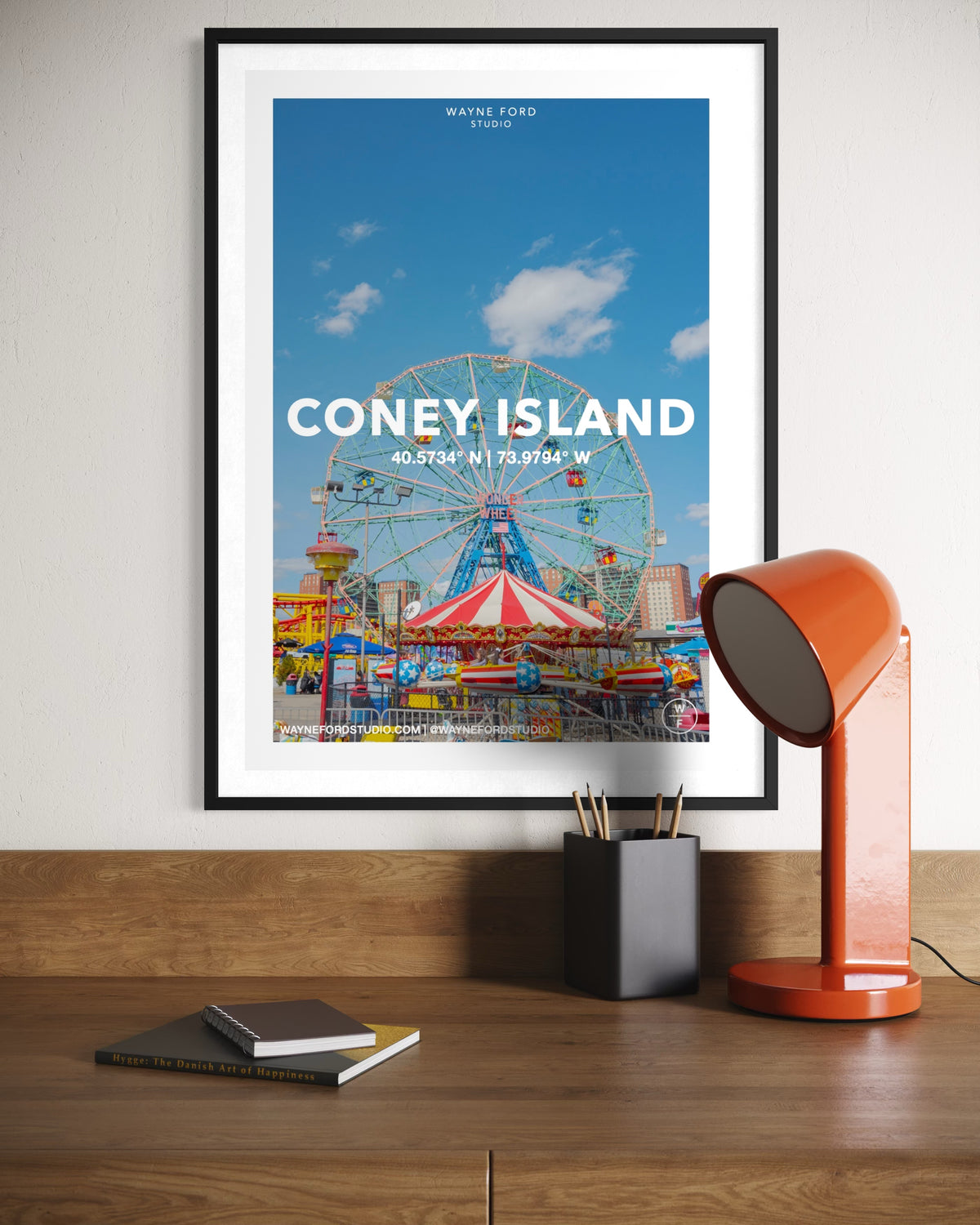 Framed Coney Island Wonder Wheel poster on desk with orange lamp, notebook, and pencil holder.