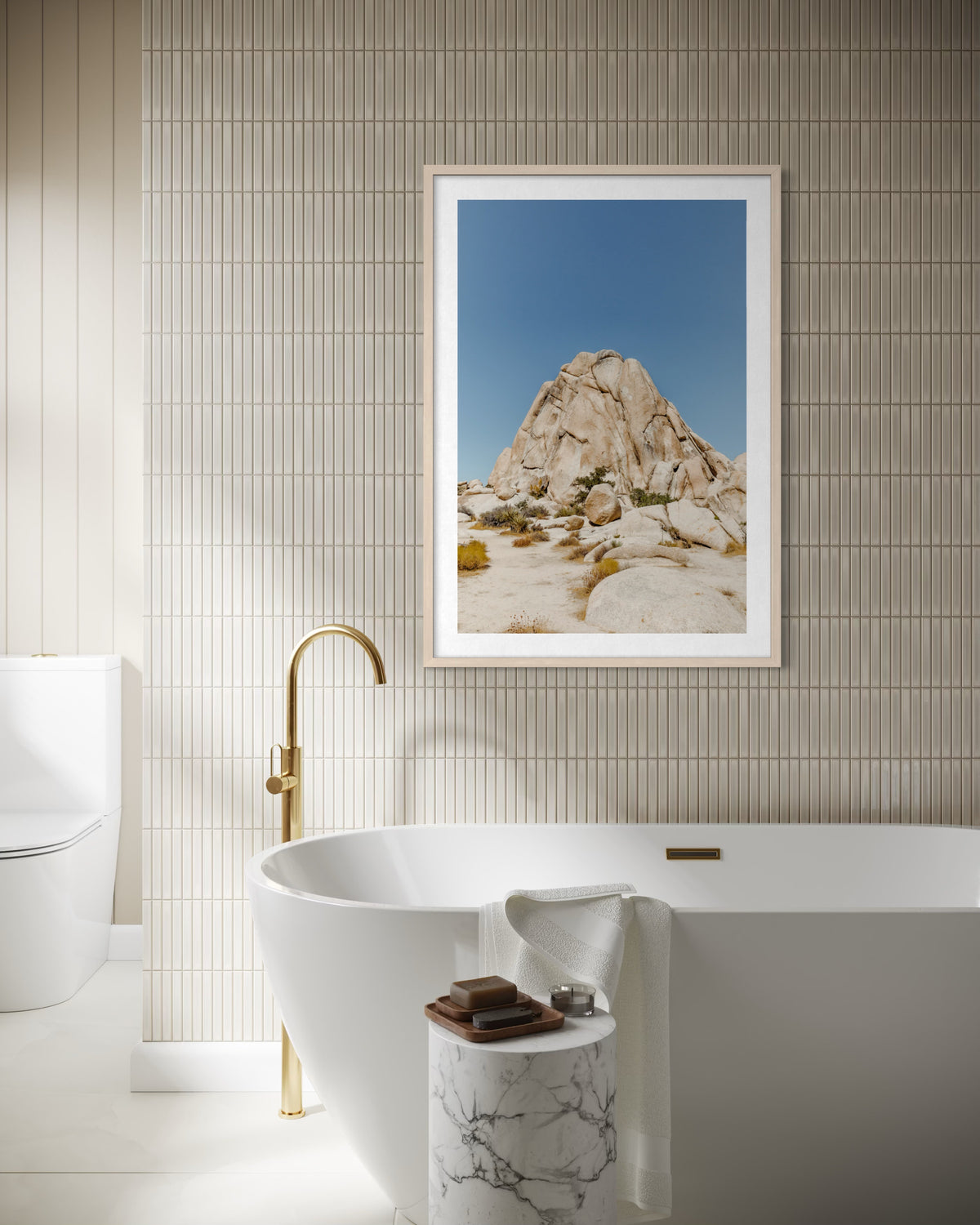 Freestanding tub, gold faucet, marble table, and Desert Form photograph on tiled bathroom wall.