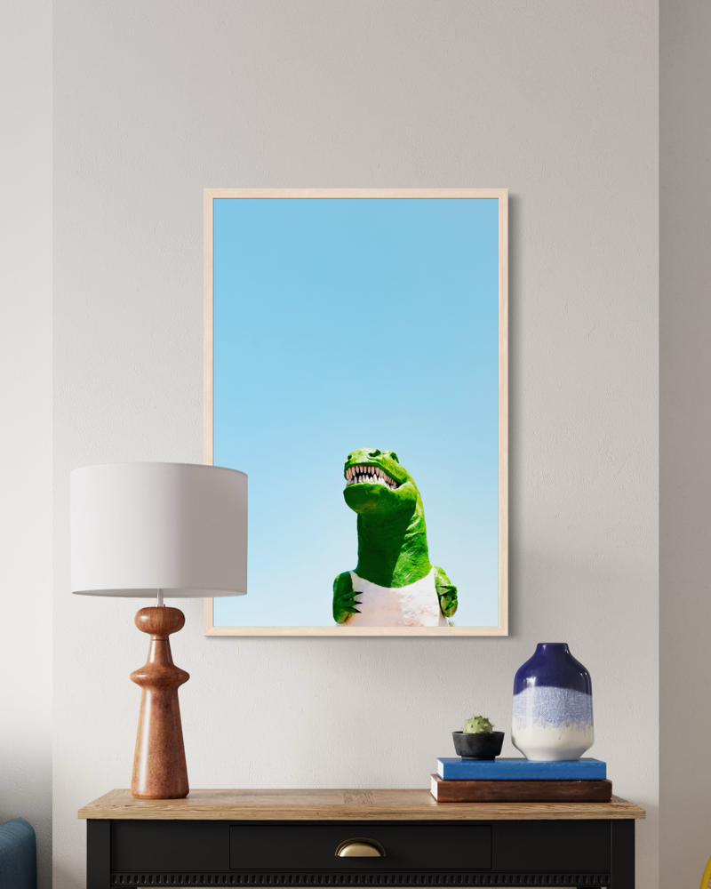 Framed print of Dino, a green dinosaur toy, under the California sky above a lamp and vases on a wooden table.