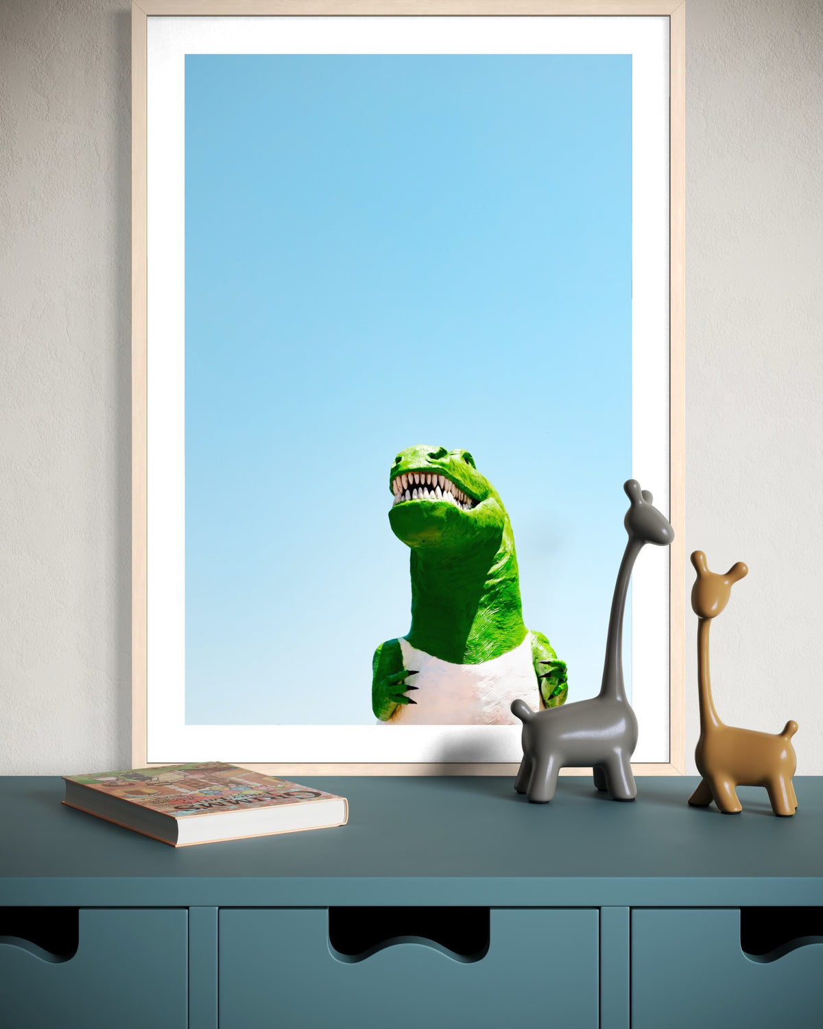 Dinosaur art print displayed beside giraffe figurines and a closed book on a shelf.
