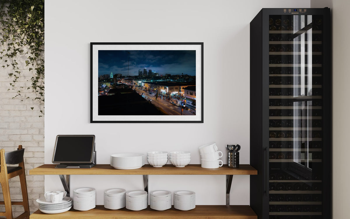 Framed Los Angeles cityscape at night displayed above dish shelves, beside a black glass wine fridge.