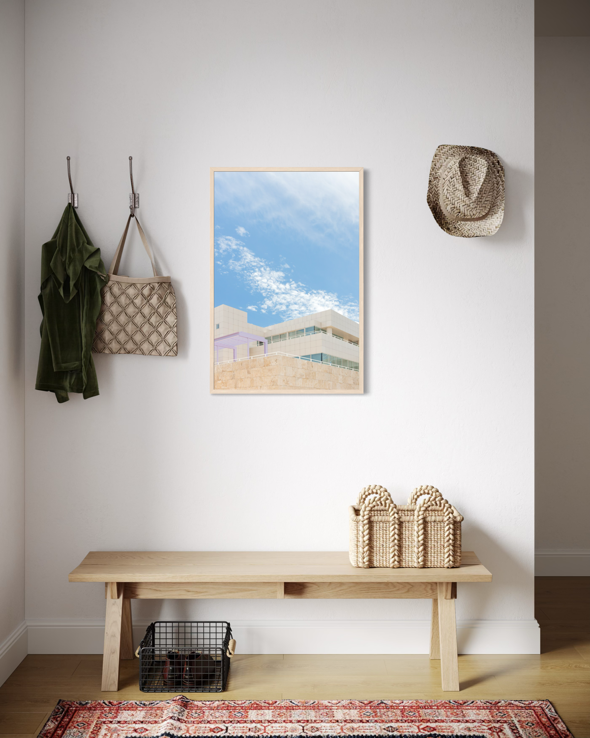 Framed Getty Center II photograph on white wall above bench with baskets, rug, green jacket, bag, and straw hat.