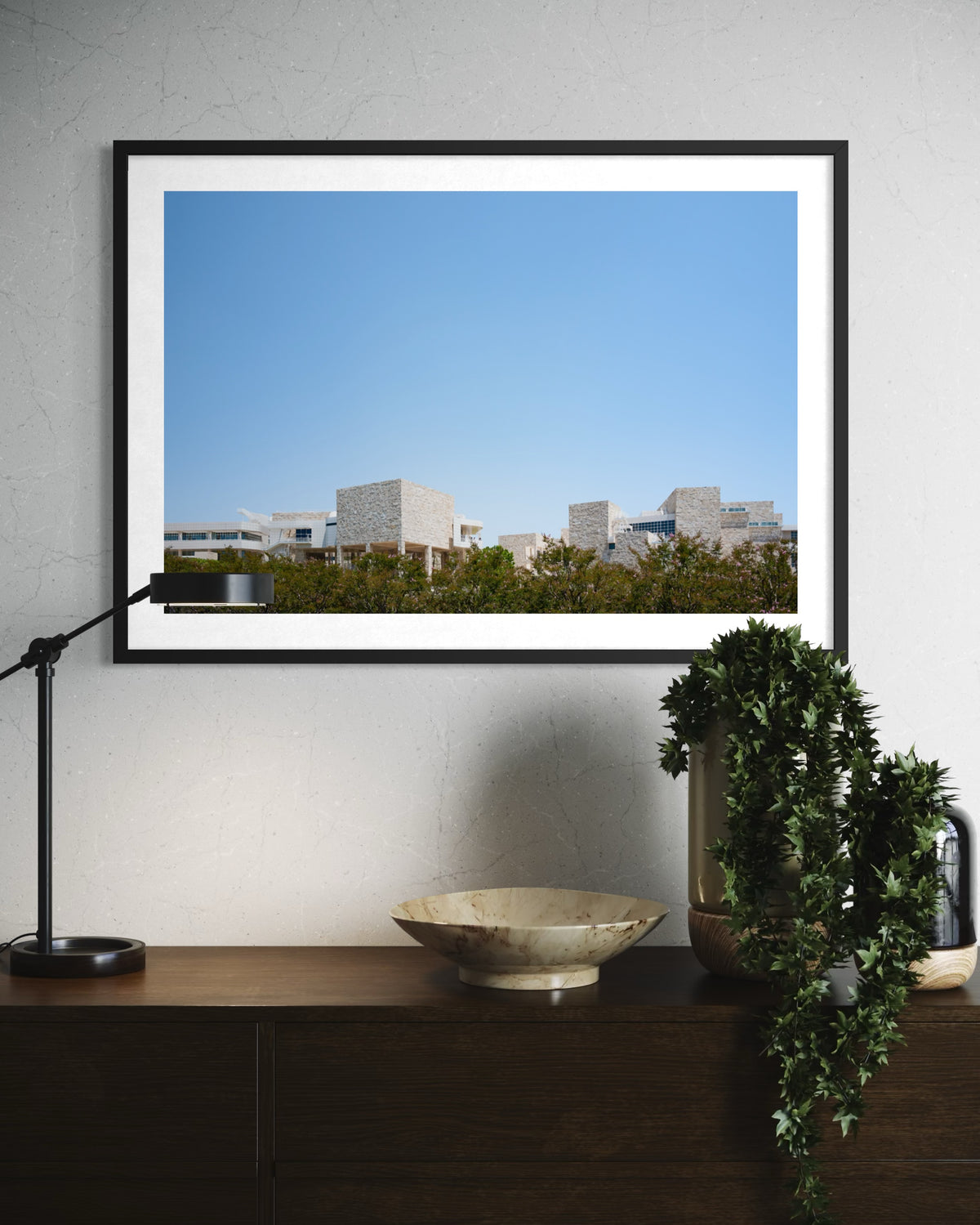 Framed Getty Center III print above a wooden console table with lamp, plant, and bowl in a room setting.