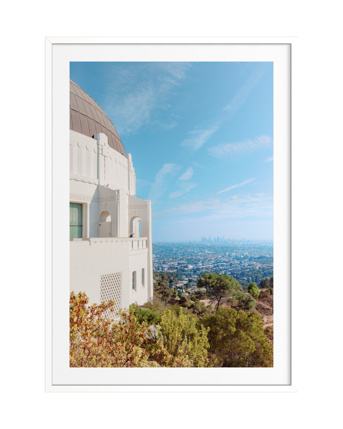 Downtown Los Angeles skyline and clear blue sky seen from Griffith Observatory, framed by surrounding trees.
