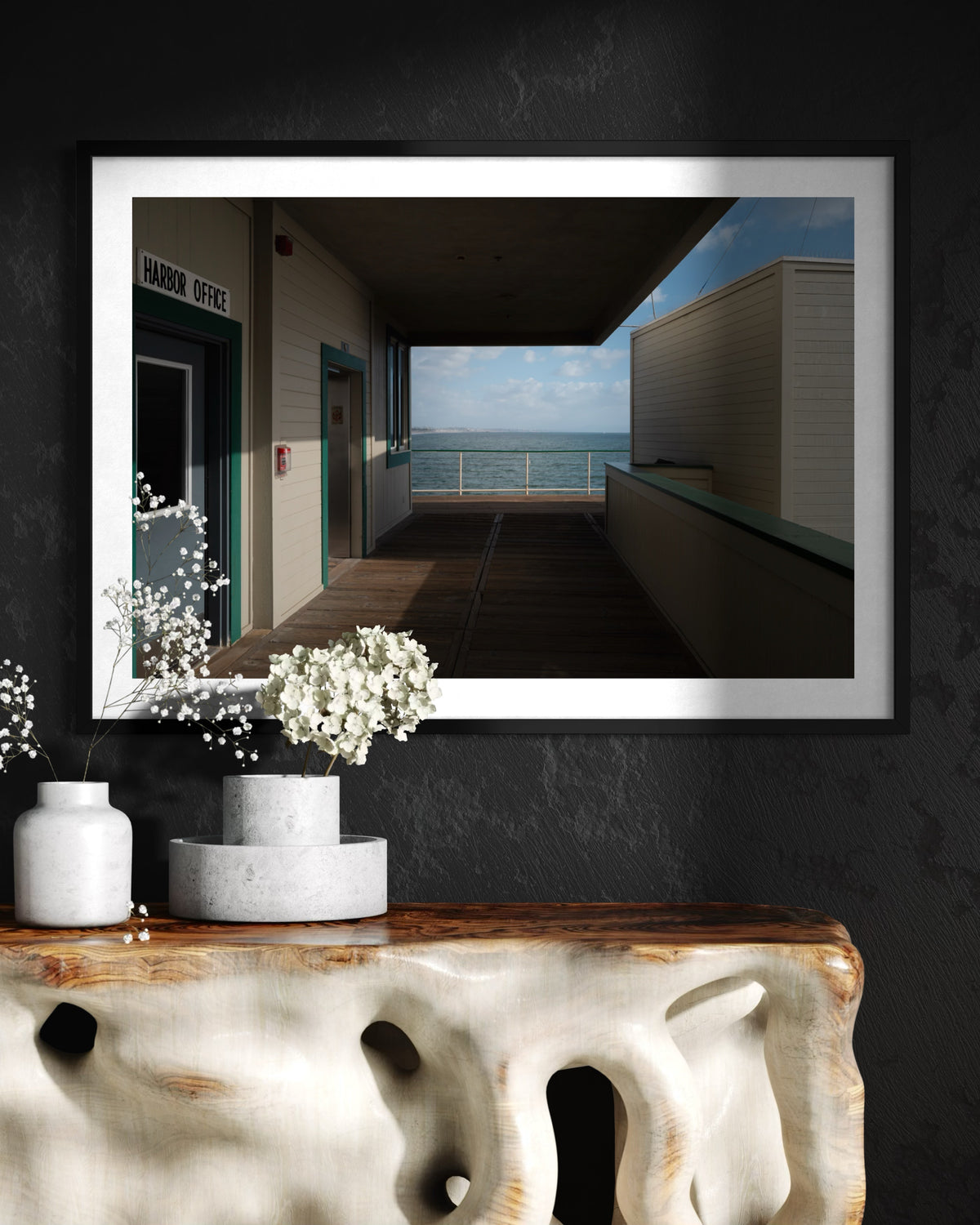 Framed Harbor Office Ocean View print above a wooden table with vases and white flowers, set against a dark wall.