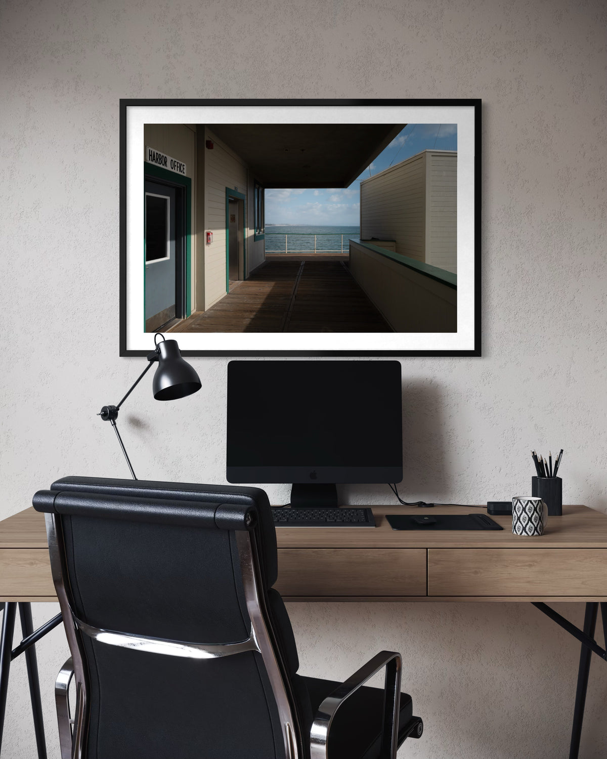 Desk with computer and lamp beneath a framed Pacific coastal walkway photograph in an office setting.