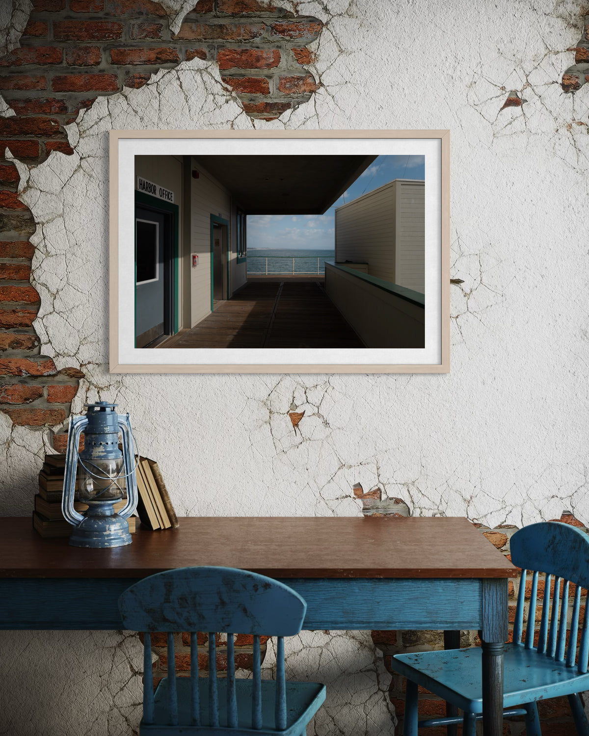 Framed print of Harbor Office and ocean view in a room above a blue table, chairs, and lantern.