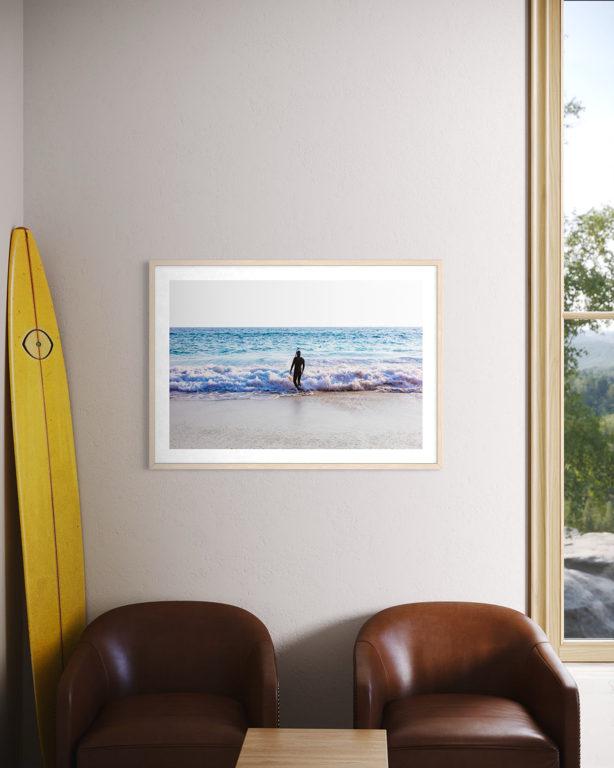 Framed “Into The Sea” beach photograph; two brown chairs, a small table, and a yellow surfboard by the wall.