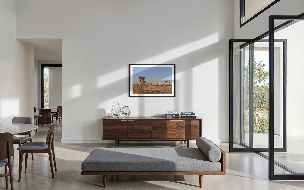 Framed Joshua Tree desert landscape at sunset above a gray chaise and wooden sideboard in a modern living room.