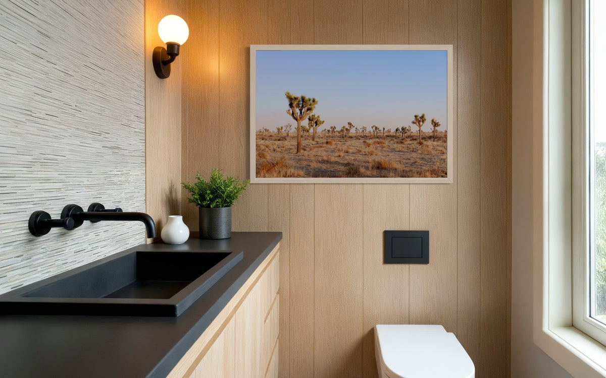 Joshua Tree landscape print hangs above a toilet in a modern bathroom with wood paneling, black sink, and potted plant.
