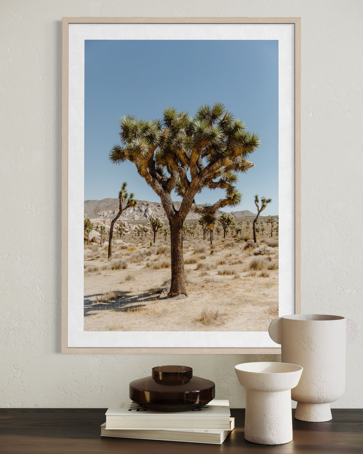 Framed landscape print of Joshua Tree National Park above stacked books and ceramics on a light-colored table.