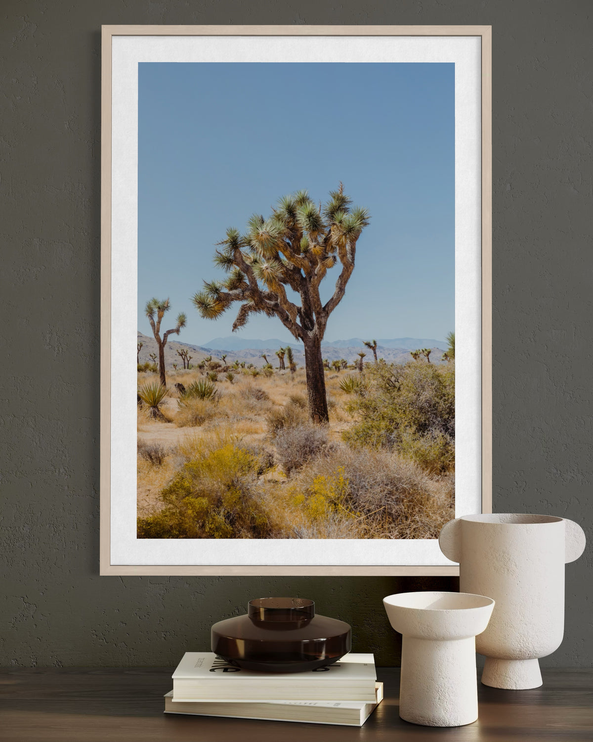 Framed photo print of a Joshua tree in the desert, displayed above a gray table with books and vases.