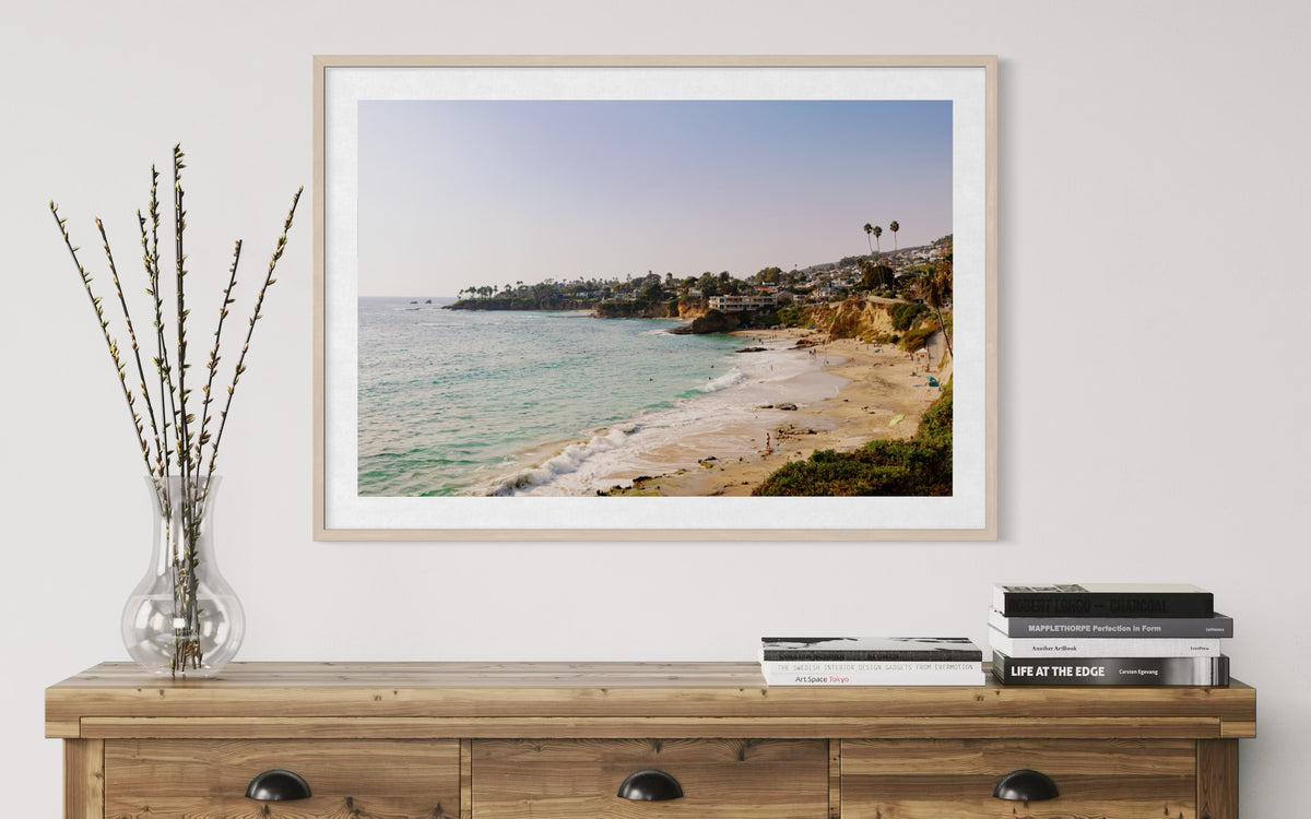 Framed photo of Laguna Beach coastline above a dresser, with branches and books in the foreground.