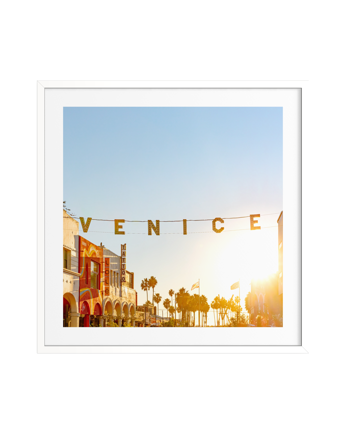 VENICE sign glowing above a sunlit Venice, CA street lined with palm trees and colorful buildings.