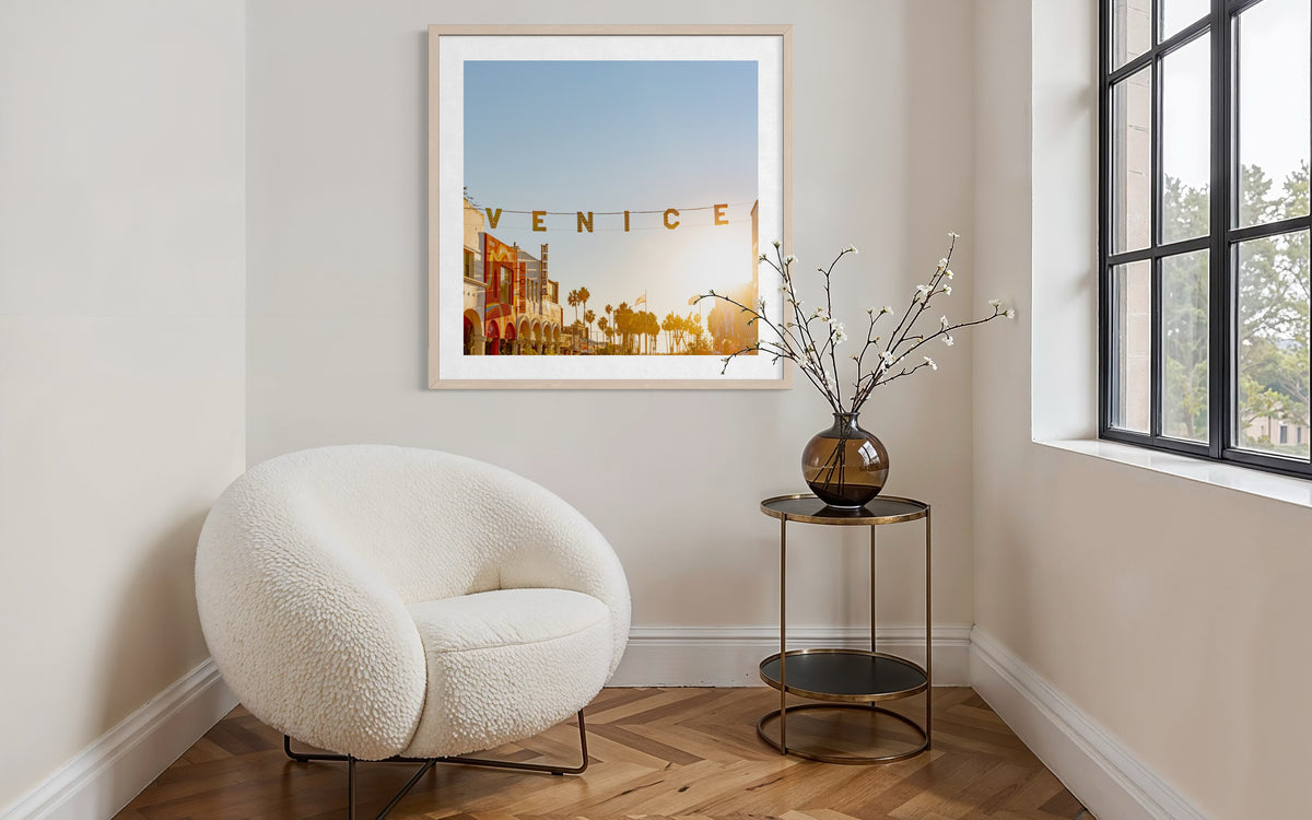 Framed Venice cityscape print glows on a wall near a window at sunset, next to a white chair and round table.