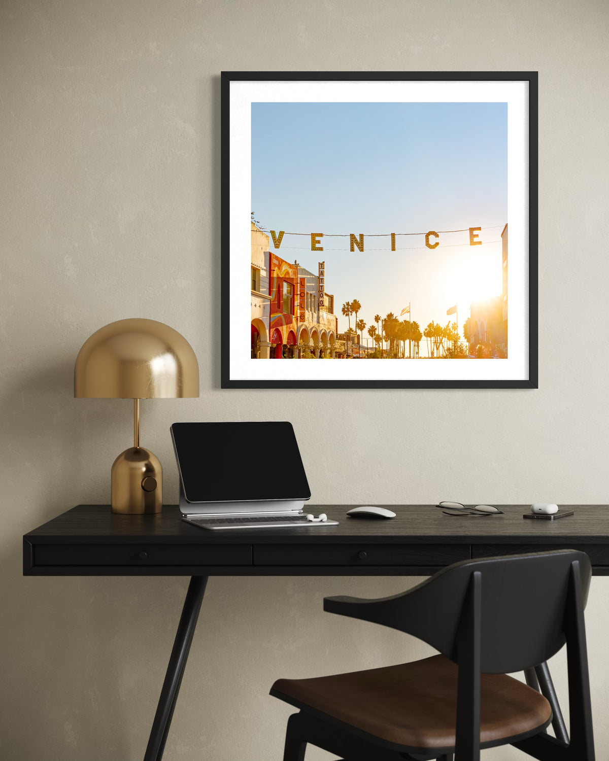 Framed print of Venice at sunset hangs above a modern desk with laptop, lamp, notebook, and black chair.