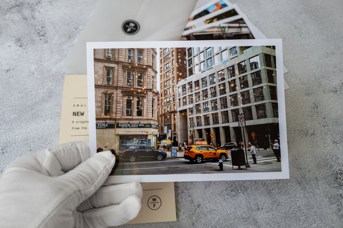 Gloved hand holding NYC Small Print Set No. 02 with street view of tall buildings and a yellow taxi.