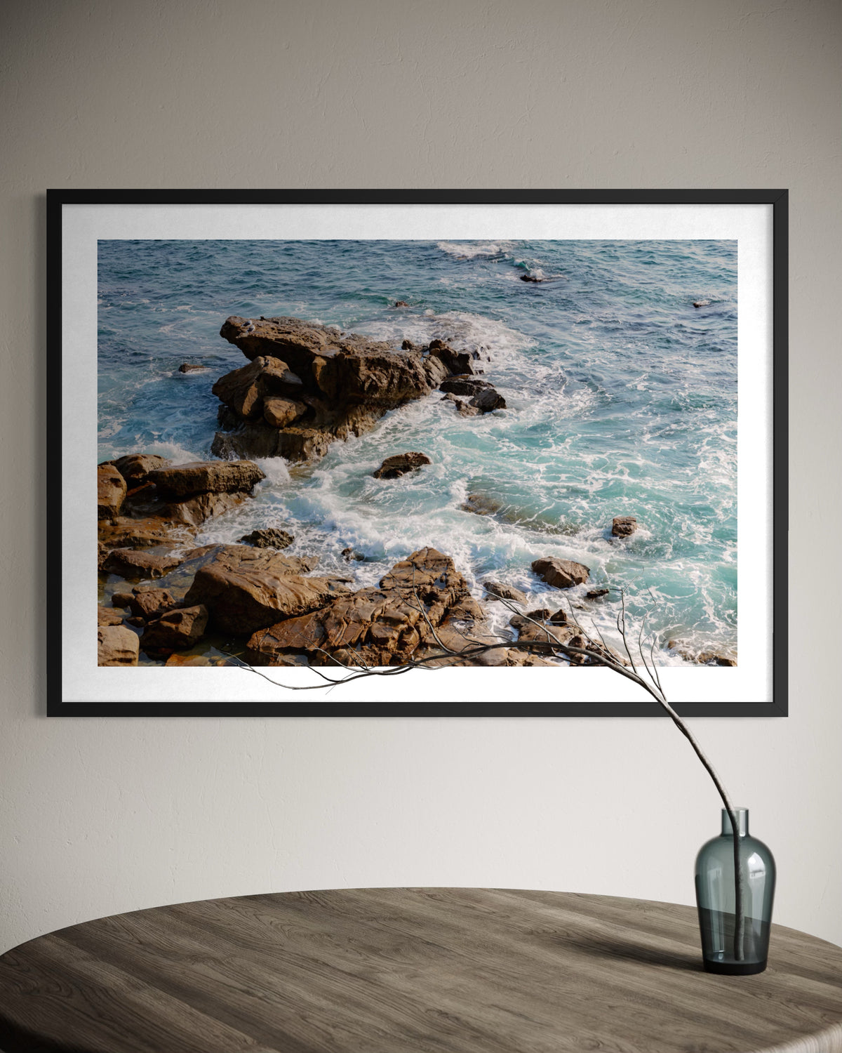 Turquoise Pacific waves crash over coastal rocks; original photo print, shown framed above a round table and vase.