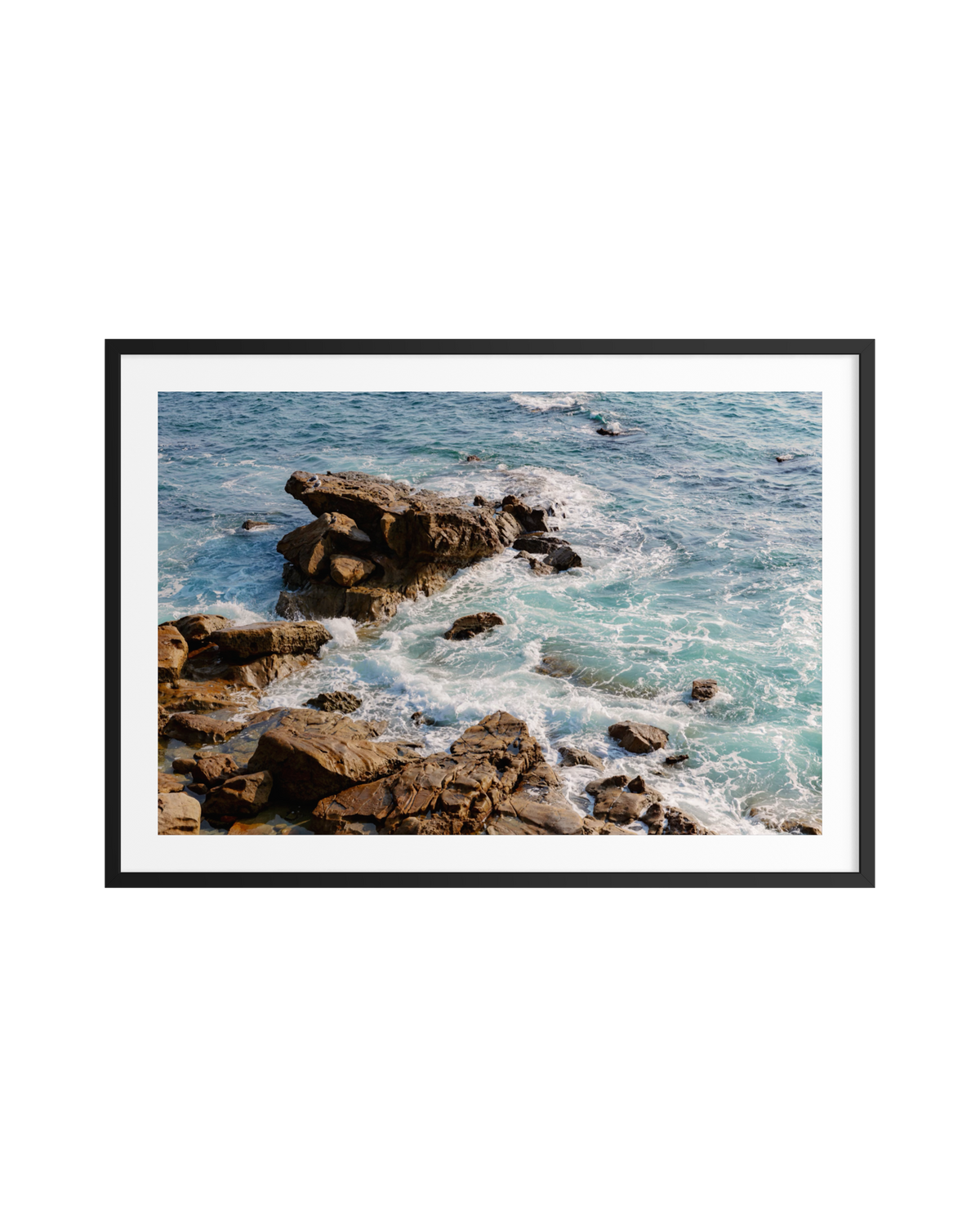 Framed print of turquoise ocean waves crashing onto rocky shore, sunlight reflecting, on a white wall.