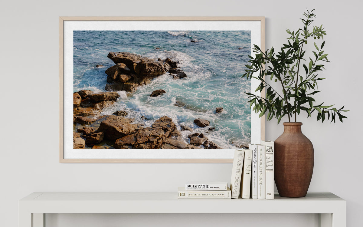 Ocean waves forming white foam, displayed in a frame above a white shelf with books and a brown vase of green branches.