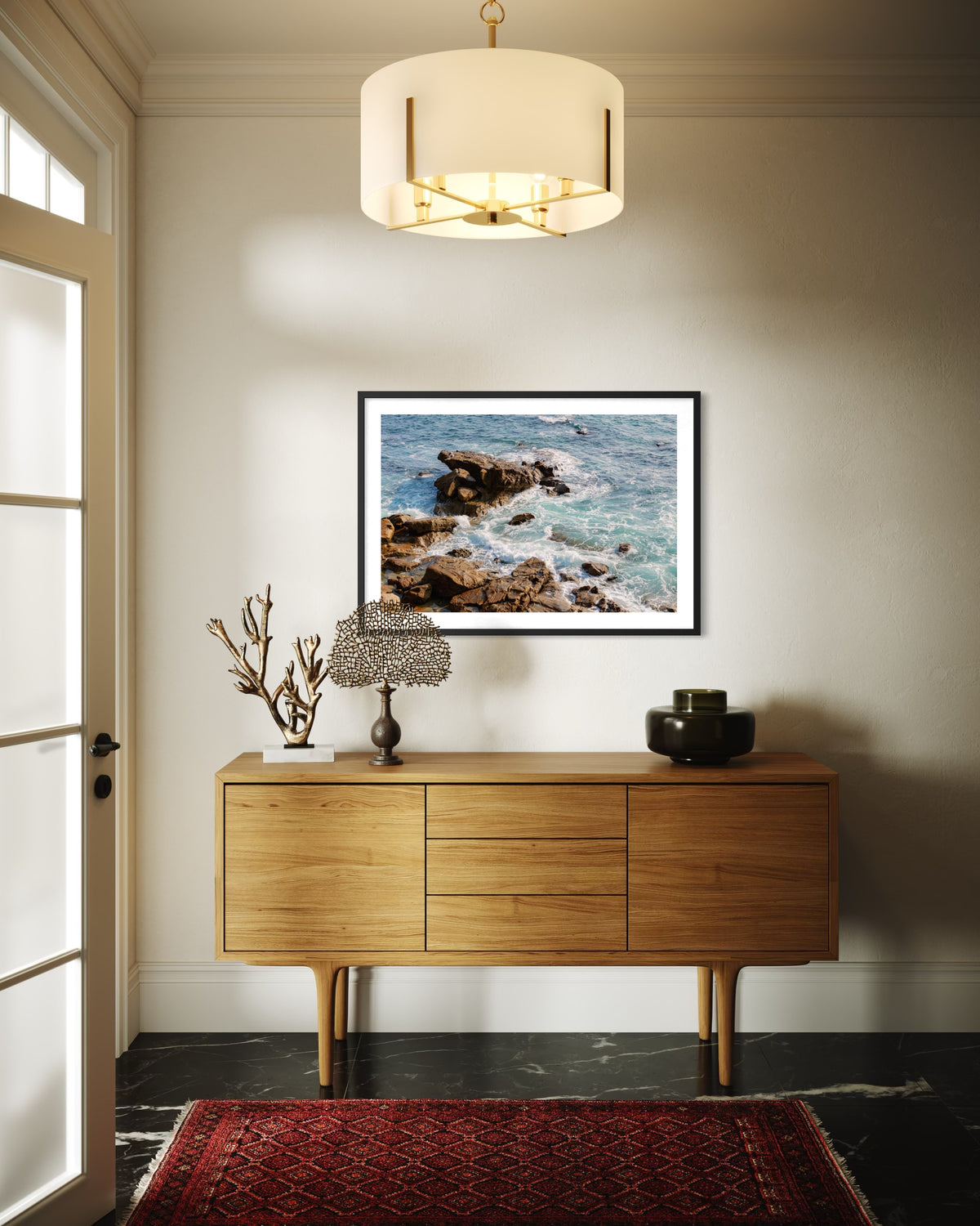 Sideboard beneath framed ocean photograph, lamp, vase, red rug, and modern chandelier in a living room setting.