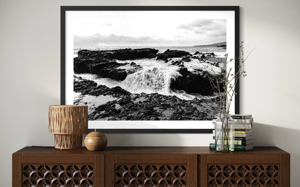 Black-and-white seascape print “Ocean Momentum” in frame, on wood cabinet with lamp, books, and vase.