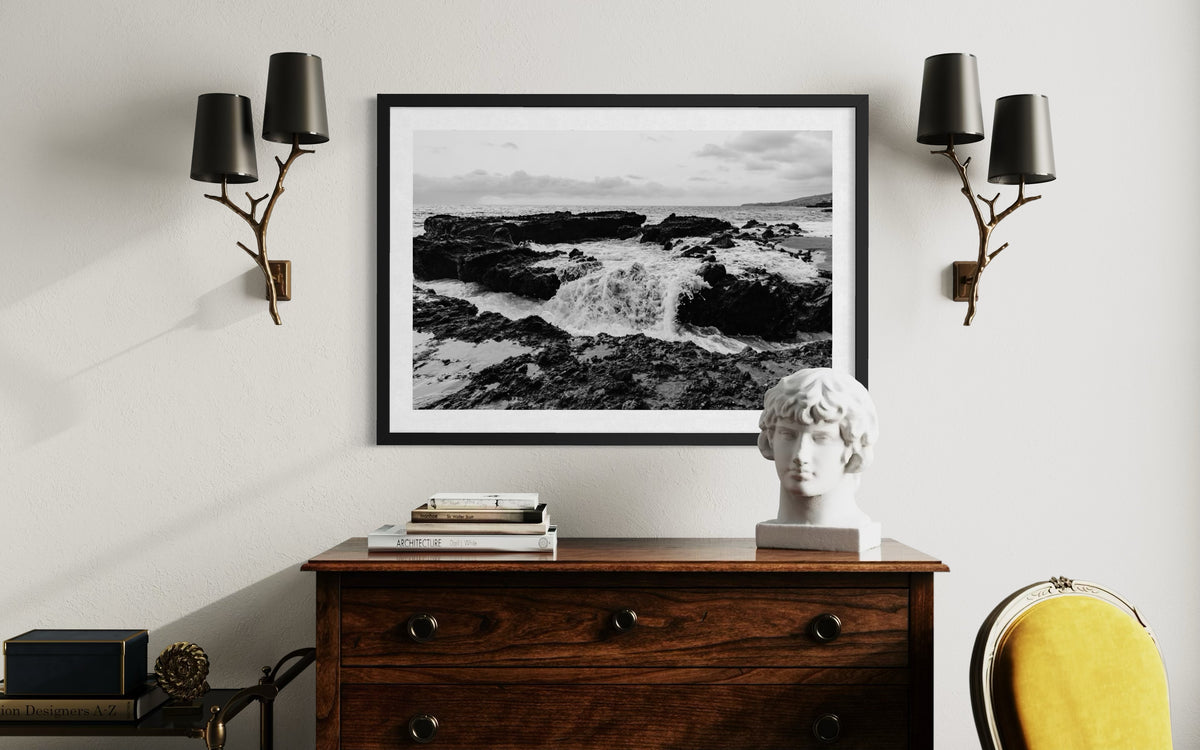 Ocean Momentum seascape print above a dresser with books, a white bust, and two black wall sconces on the wall.