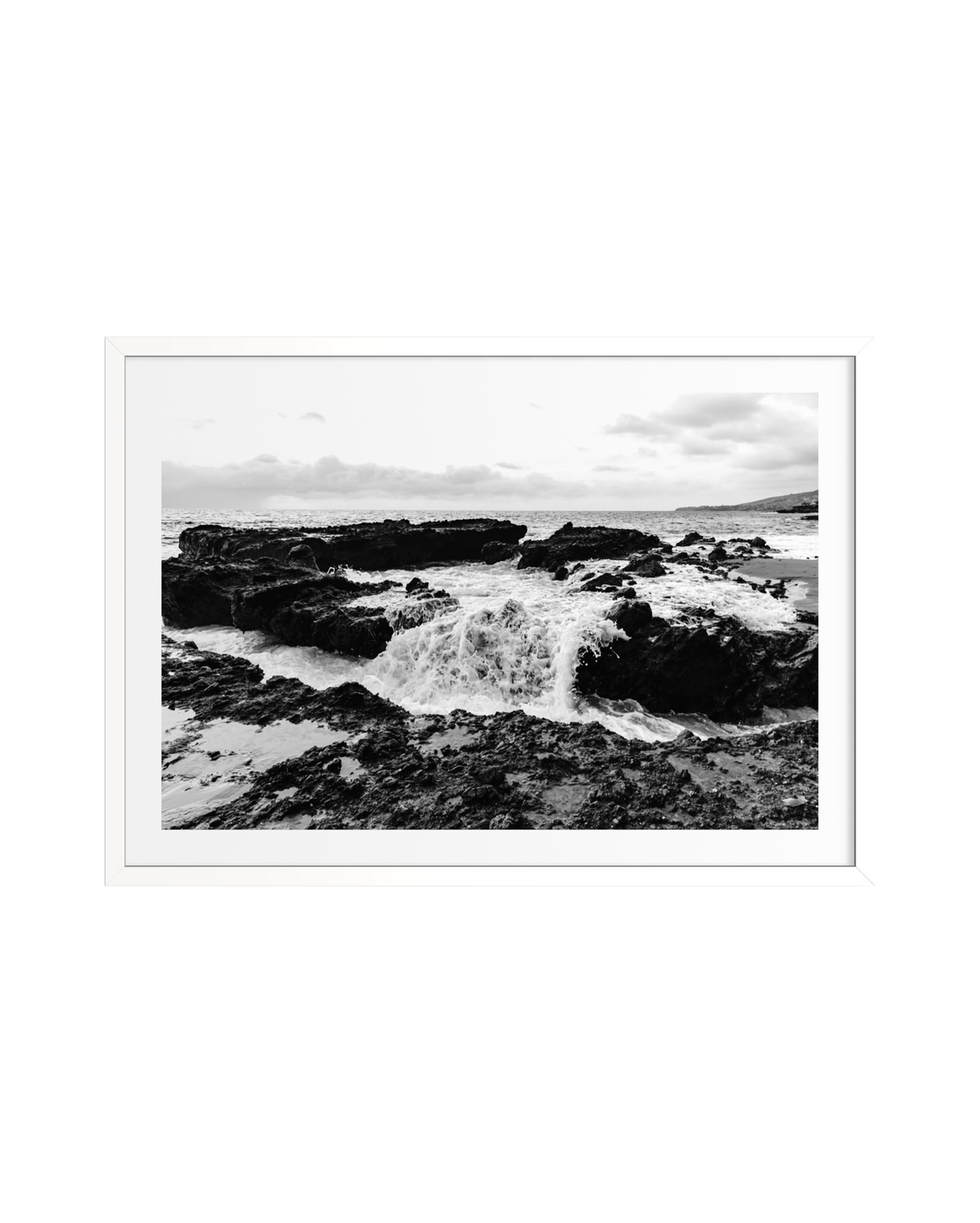 Waves crash against rocks beneath cloudy skies, captured in black and white and framed with a white border.