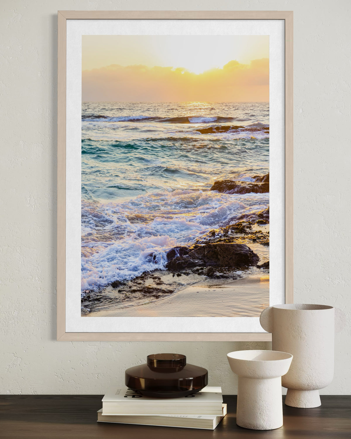 Framed photo print of a rocky beach at dusk, placed above books and ceramics on a tabletop.