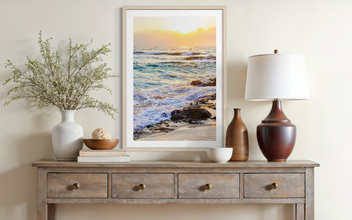 Framed Ocean Sunset photo on a light wall above a wooden table with vases and a lamp.