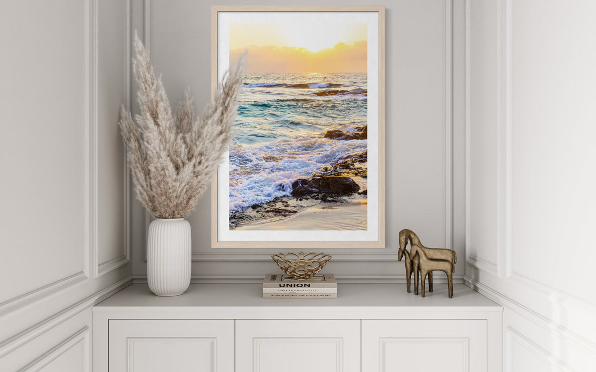 Framed ocean sunset print on white cabinet with pampas grass, horse figurines, and stacked books.