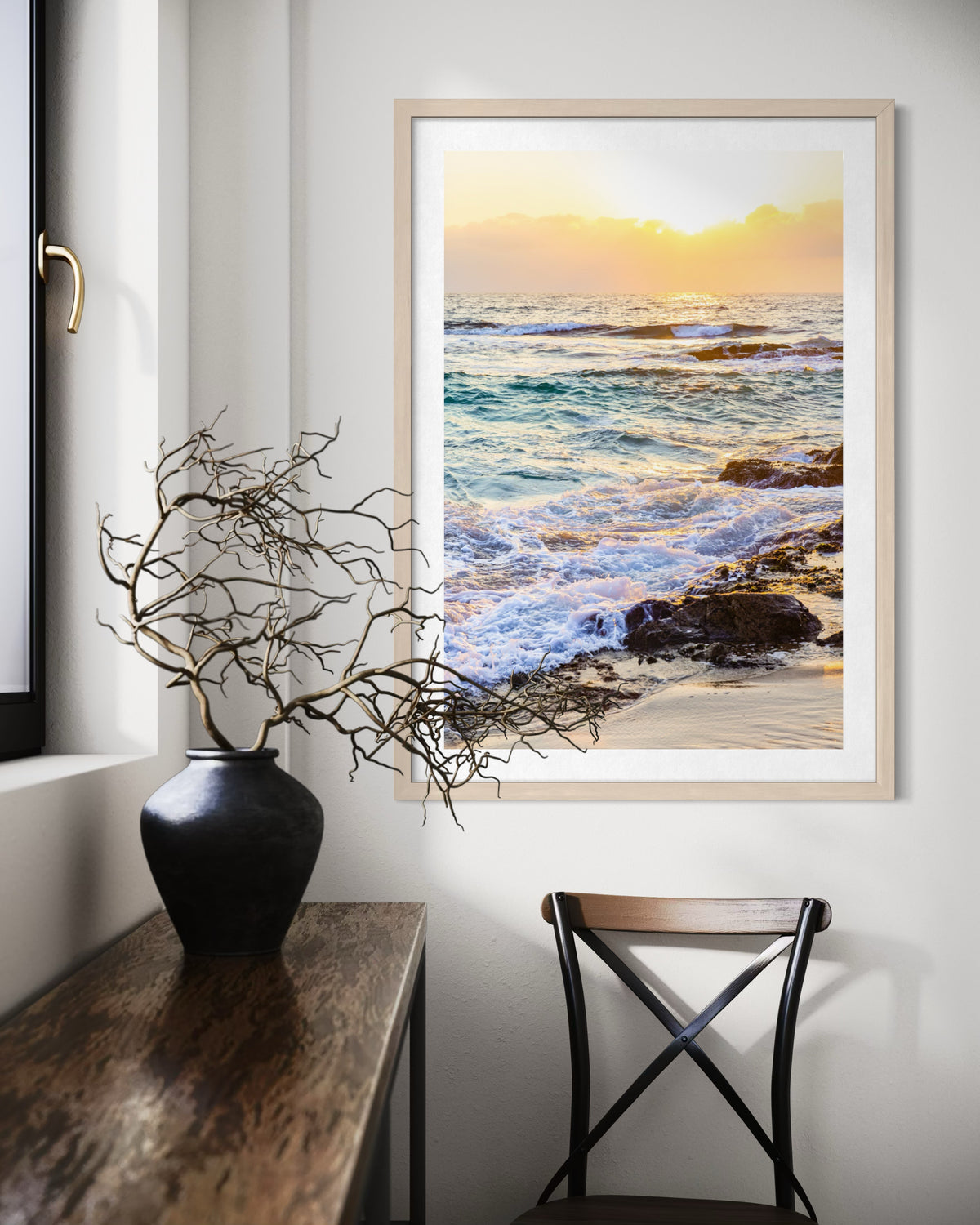 Framed “Ocean Sunset” print above a chair, beside a table with a vase of dried branches in the room.