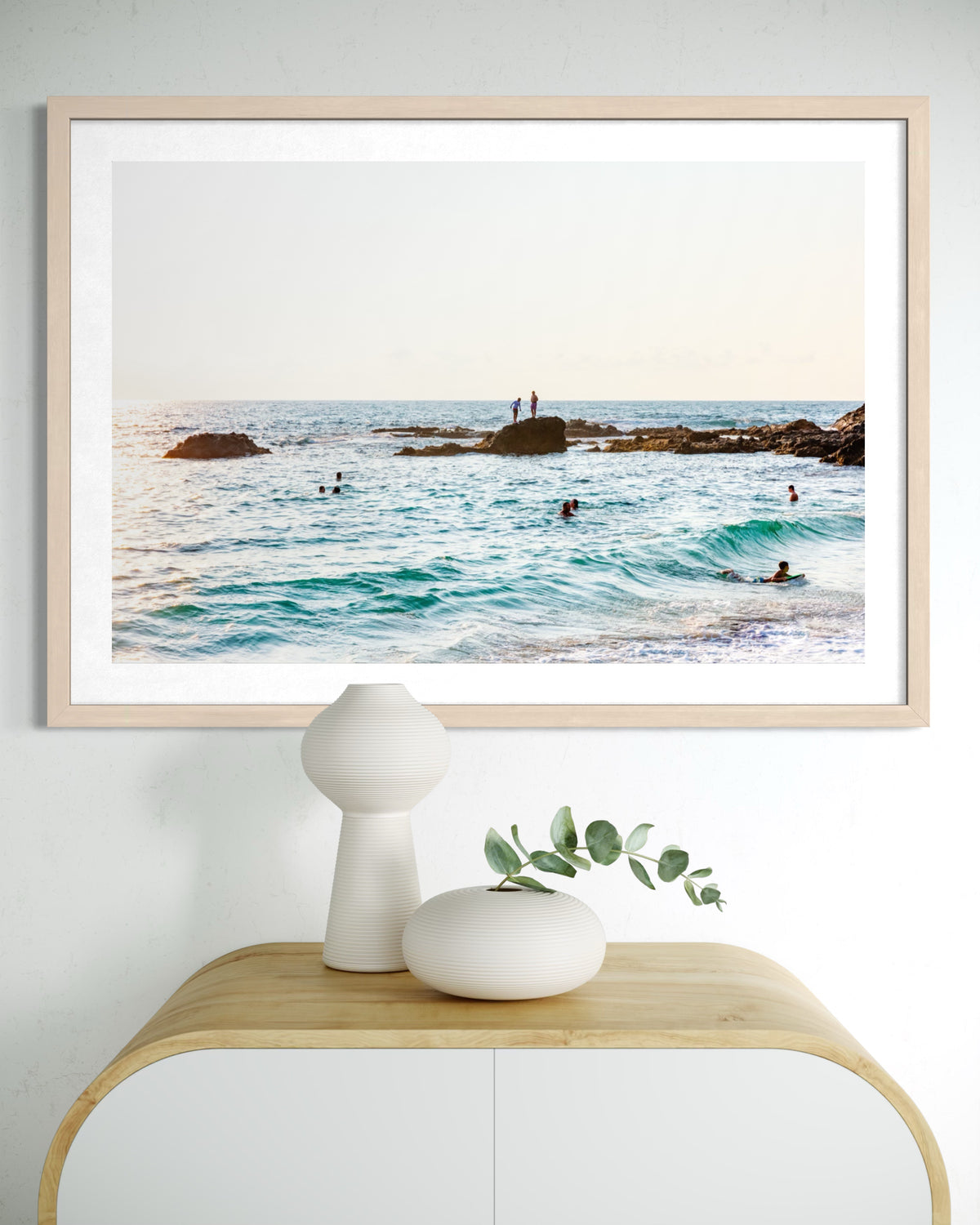 Framed print of swimmers and rock climbers at Laguna Beach, displayed above a light wood table with vases.