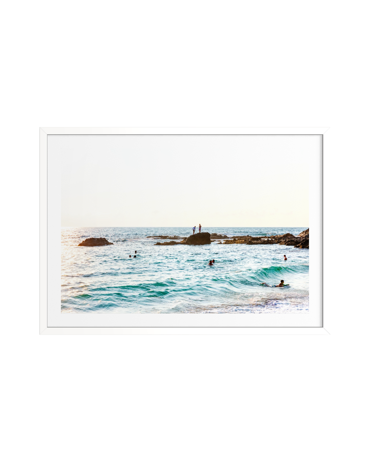 Two people sit on rocks overlooking swimmers in Pacific waves at Laguna Beach, California, under a clear sky.