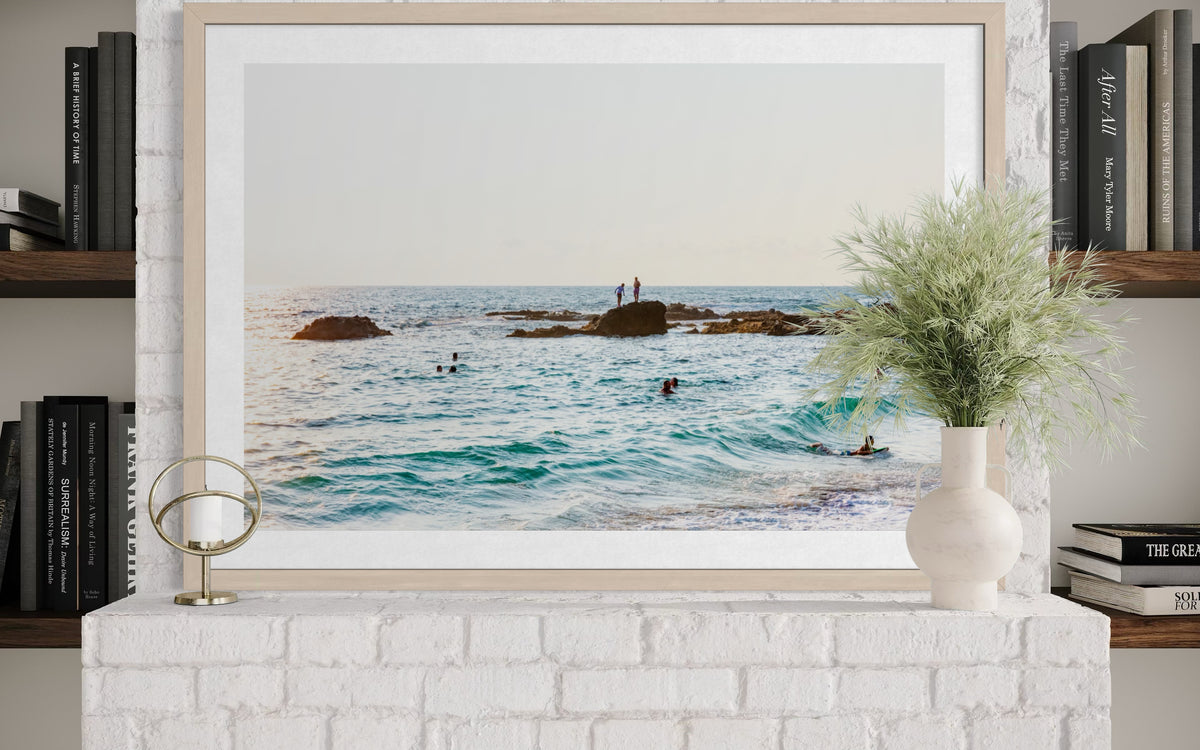 Framed photo print of swimmers in the ocean displayed on a white brick mantel with books and a small potted plant.