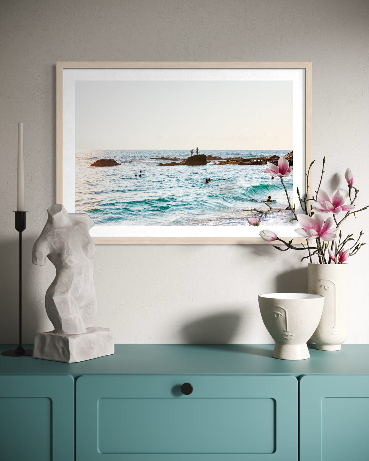 Above a teal cabinet, an ocean swim photograph with abstract sculpture, magnolias, and two face vases.