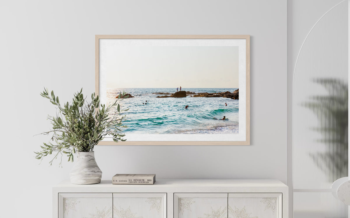 Framed “Ocean Swim” photograph of swimmers in blue water, above a white cabinet with marble vase and books.