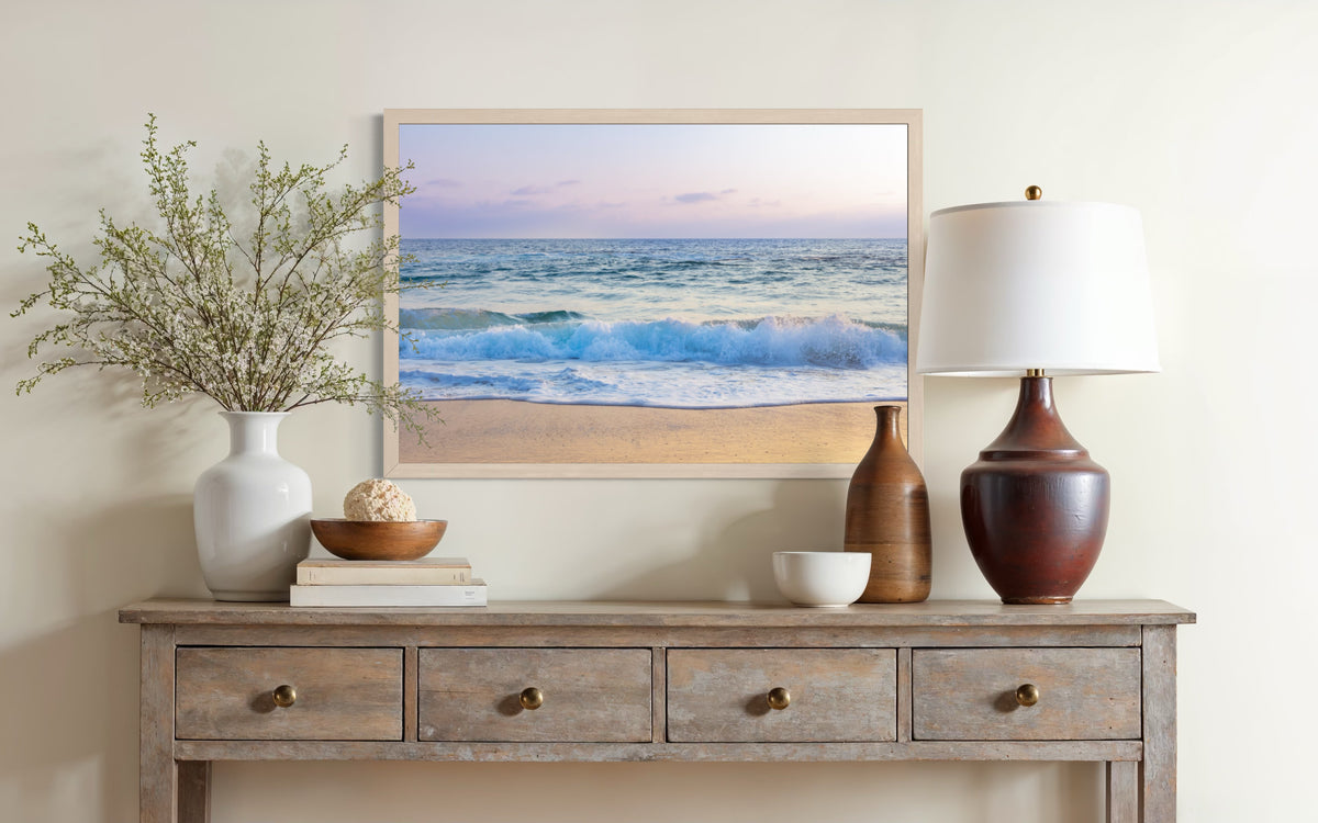 Framed “Ocean Waves” print above a lamp and decor on a wooden table in a softly lit room.