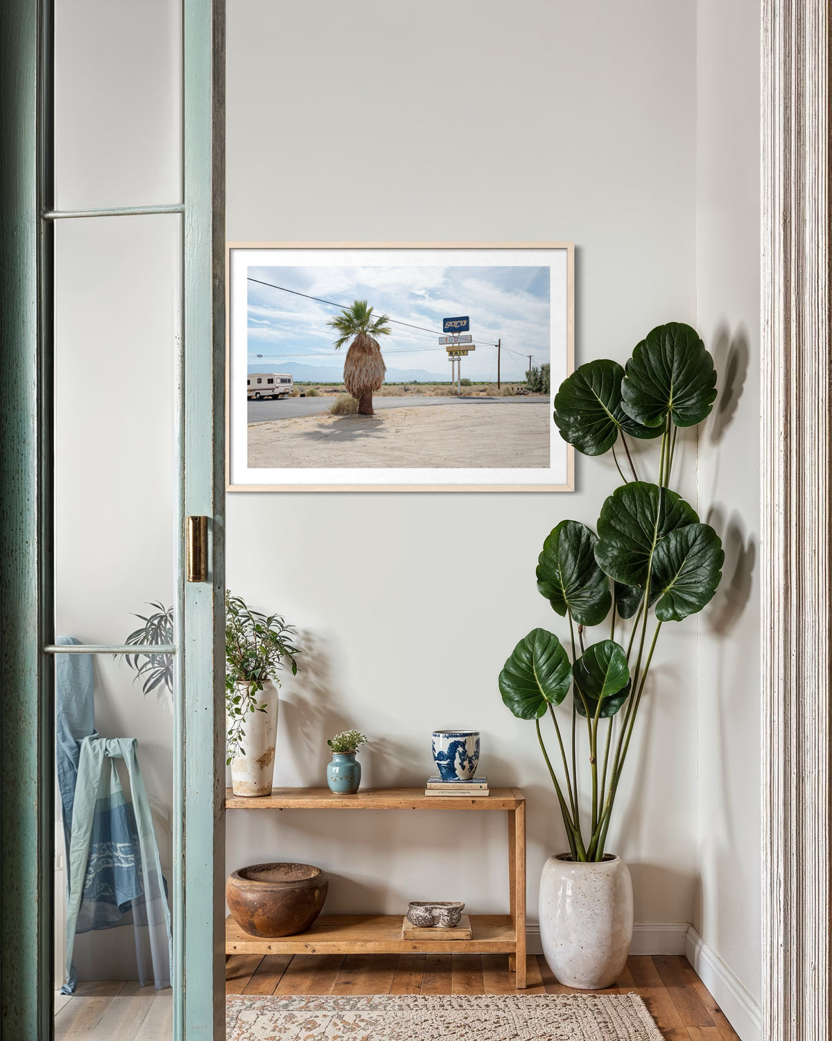 Palm Desert roadside print displayed above a wooden shelf with pottery and a leafy plant in a white pot.