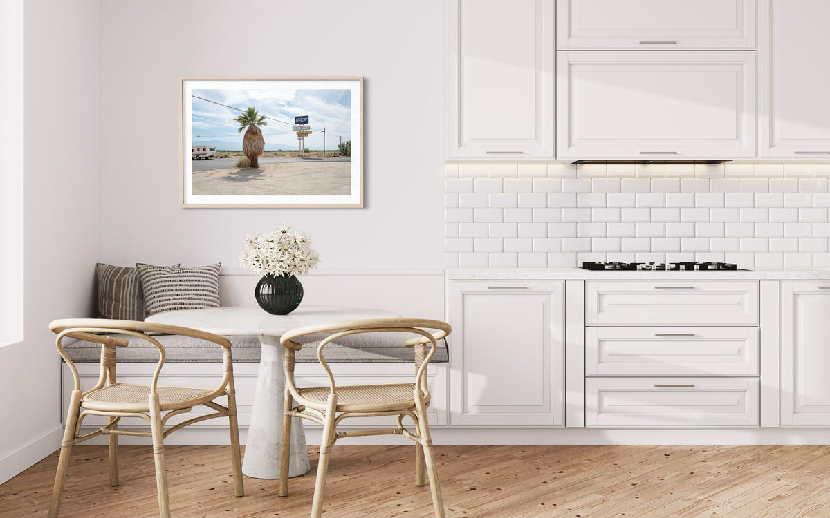 Framed “Palm Desert Roadside” photo on wall in white kitchen with round table and two chairs.