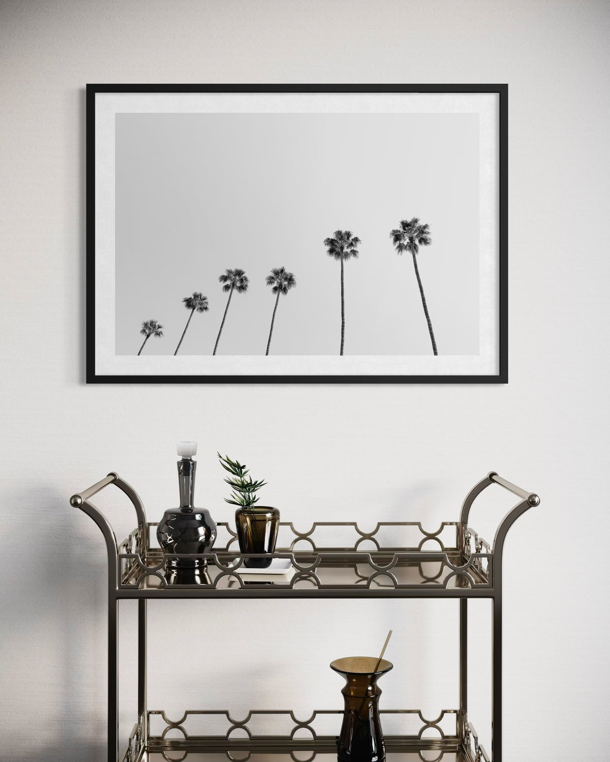 Artwork featuring palm trees hangs above a bar cart with glassware, bottles, and a potted plant.
