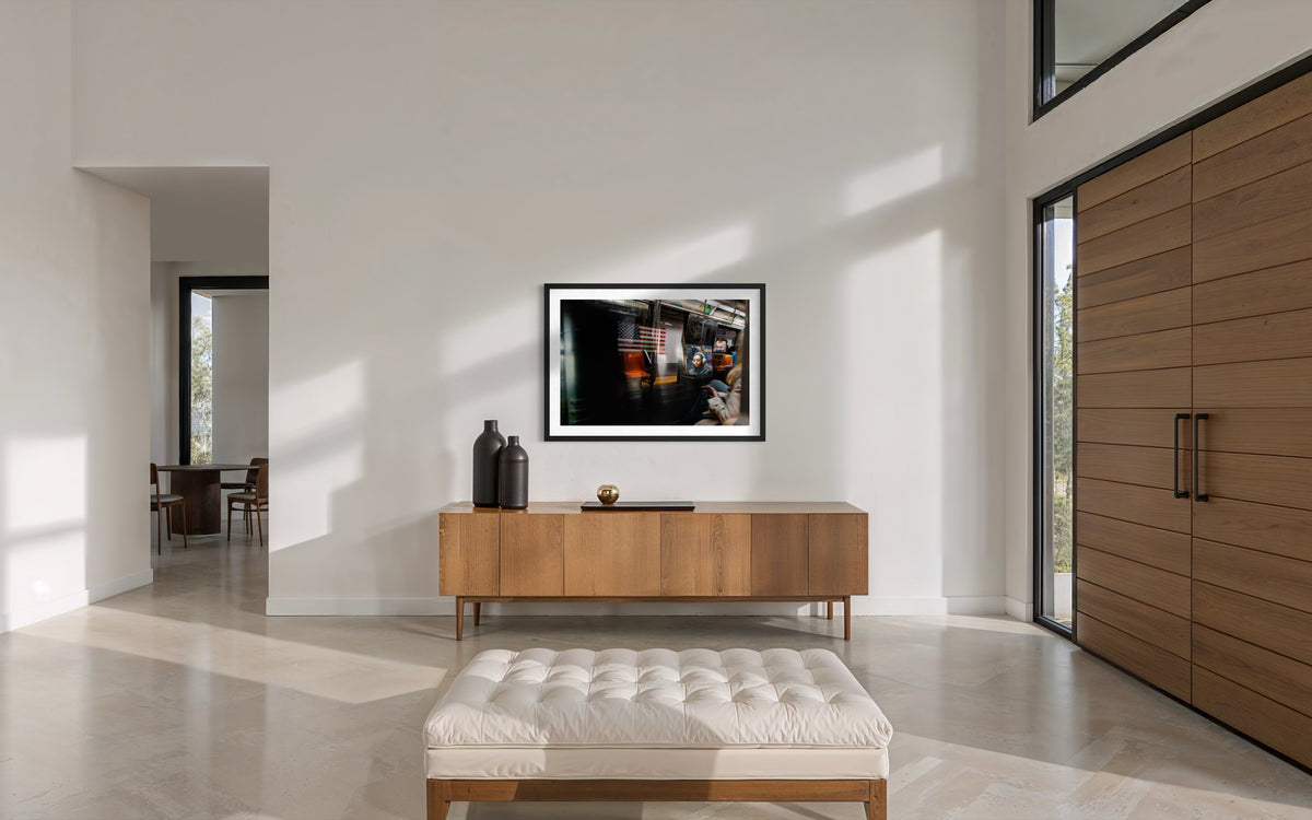 Framed “Passing Train, New York City” photo above wood sideboard in a white living room with large windows.