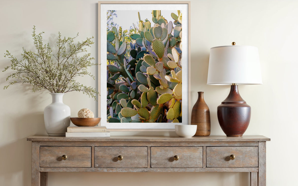 Framed “Prickly Pear” cactus photo displayed on a wooden console with decor and lamp, set against a light wall.