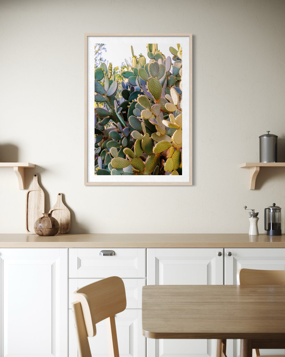 Framed prickly pear cactus print displayed on a beige kitchen wall above white cabinets and wooden kitchenware.