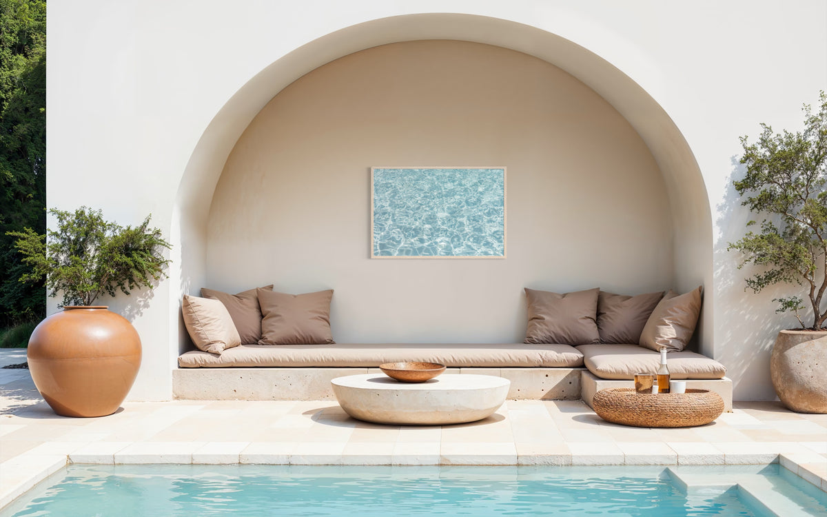 Modern outdoor lounge with tan cushions, round tables, and poolside water views beside an arched wall.