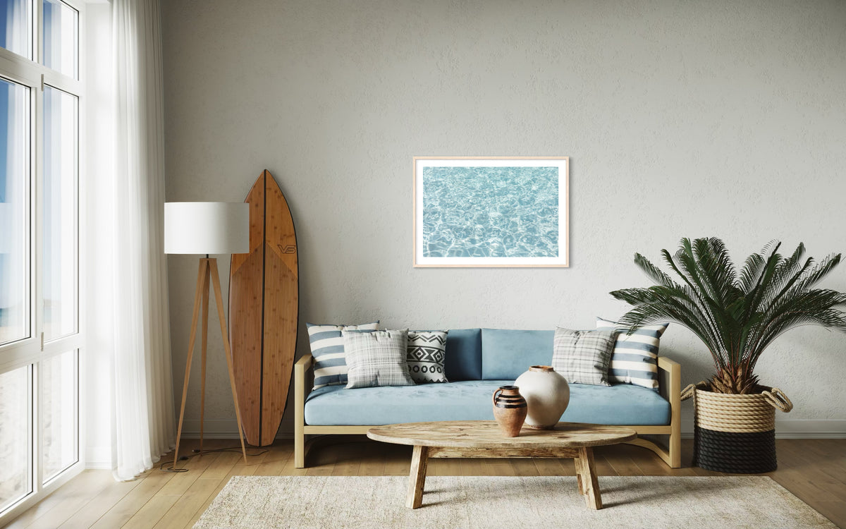 Blue couch and surfboard in bright living room, with “Refresh II” beach print framed on the wall.